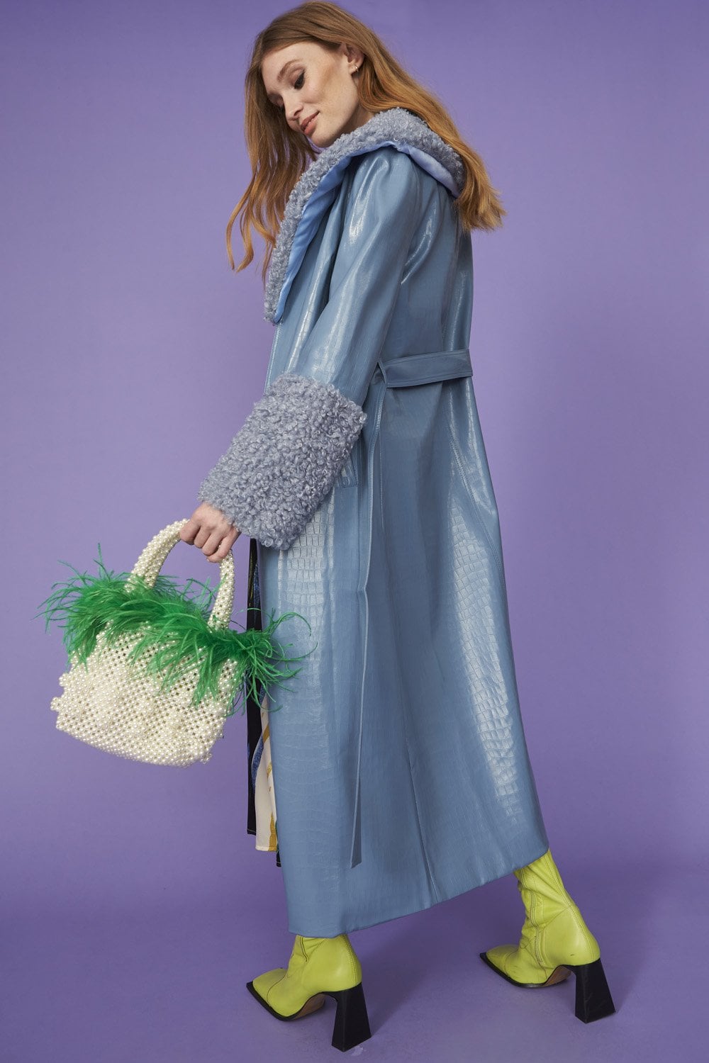 A woman in a blue faux suede coat with detachable faux shearling cuffs stands against a purple background, holding a white beaded bag adorned with green feathers and wearing yellow boots.