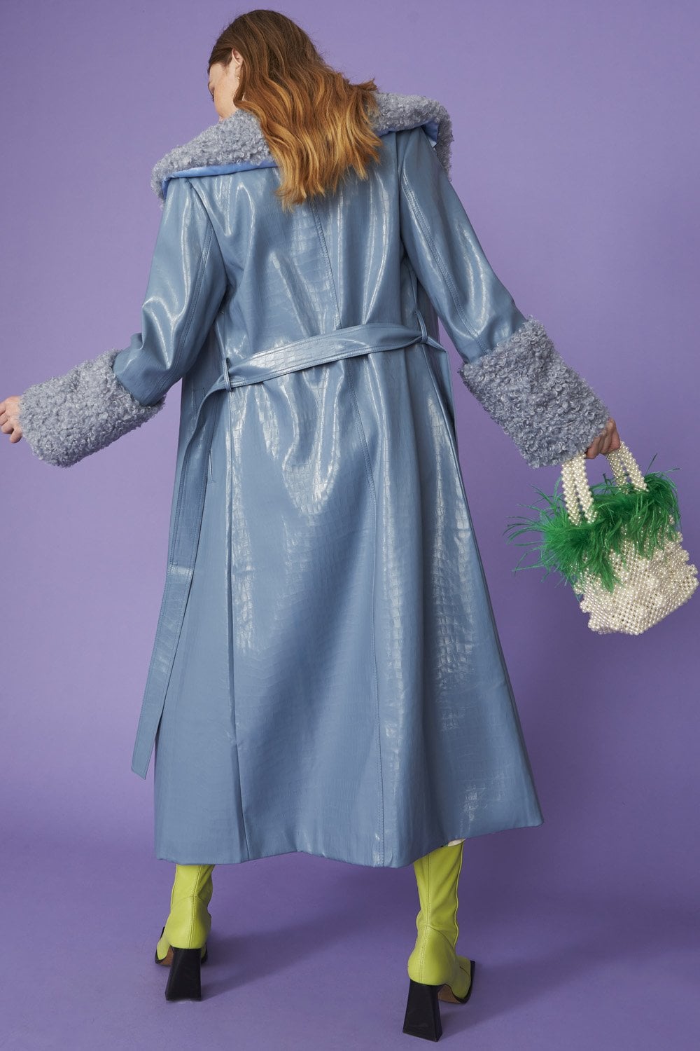 A person in a shiny blue faux suede coat with detachable faux shearling cuffs, yellow-green tights, black-heeled shoes, and a beaded bag adorned with green feathers poses against a purple background.