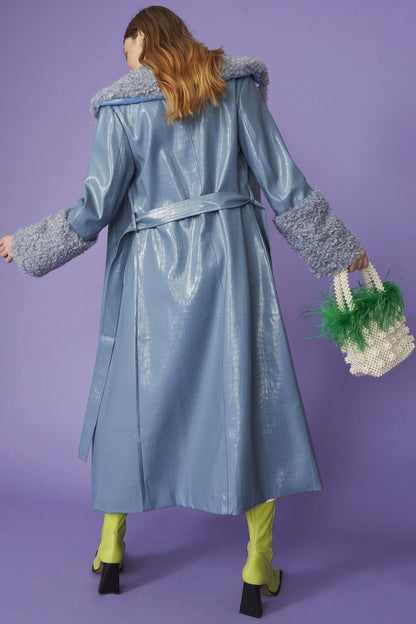 A person in a shiny blue faux suede coat with detachable faux shearling cuffs, yellow-green tights, black-heeled shoes, and a beaded bag adorned with green feathers poses against a purple background.