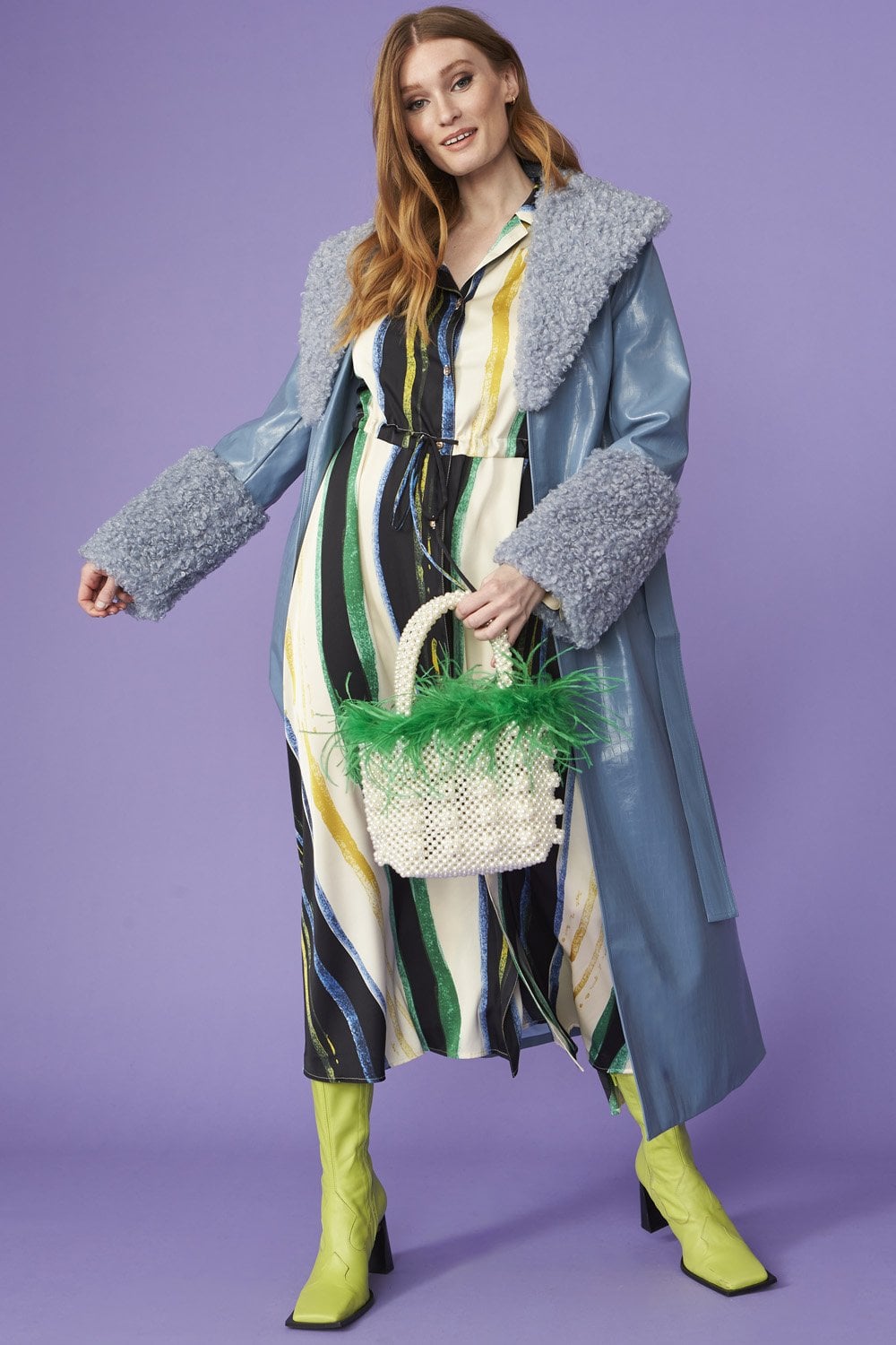 Woman poses in a blue faux suede coat with detachable faux shearling cuffs, striped dress, neon green boots, and holds a woven bag with green feather details against a purple background—ideal for winter layering.