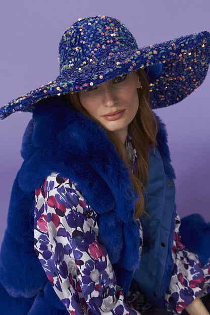 Woman wearing a wide-brimmed, blue sequin flapper hat, a blue fur gilet, and a floral patterned blouse, seated in front of a purple background for a touch of vintage glamour.