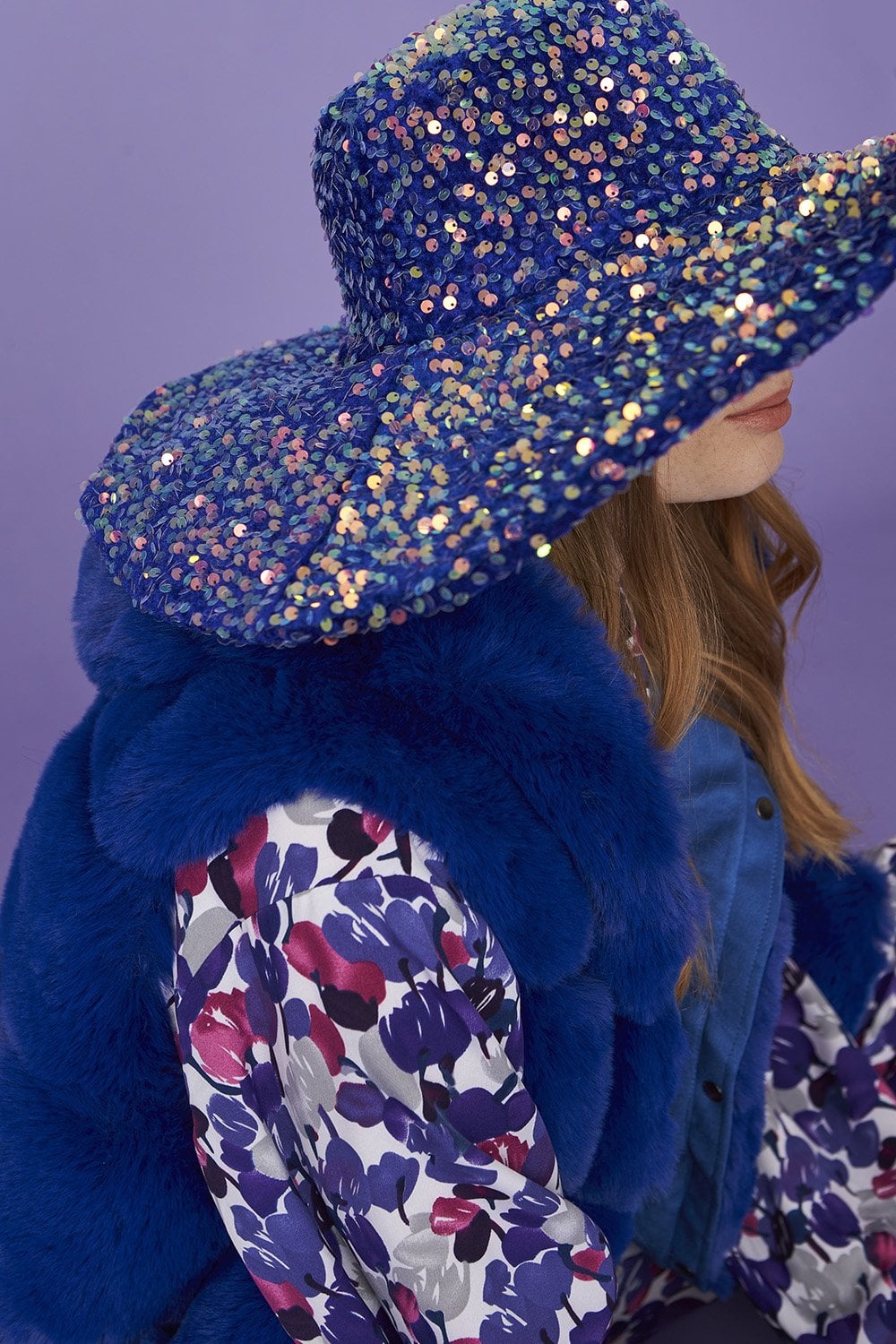 A person wearing a vintage glamour blue sequin flapper hat, a blue fur gilet, and a floral patterned shirt set against a purple background.