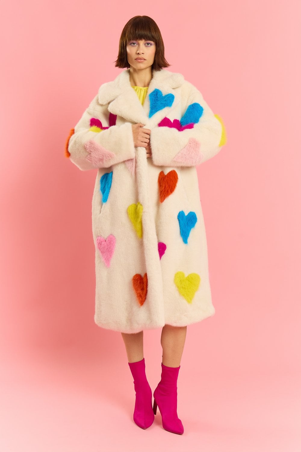 A woman stands against a pink background wearing a long, white sustainable faux fur coat adorned with colourful heart shapes and fuchsia ankle boots.