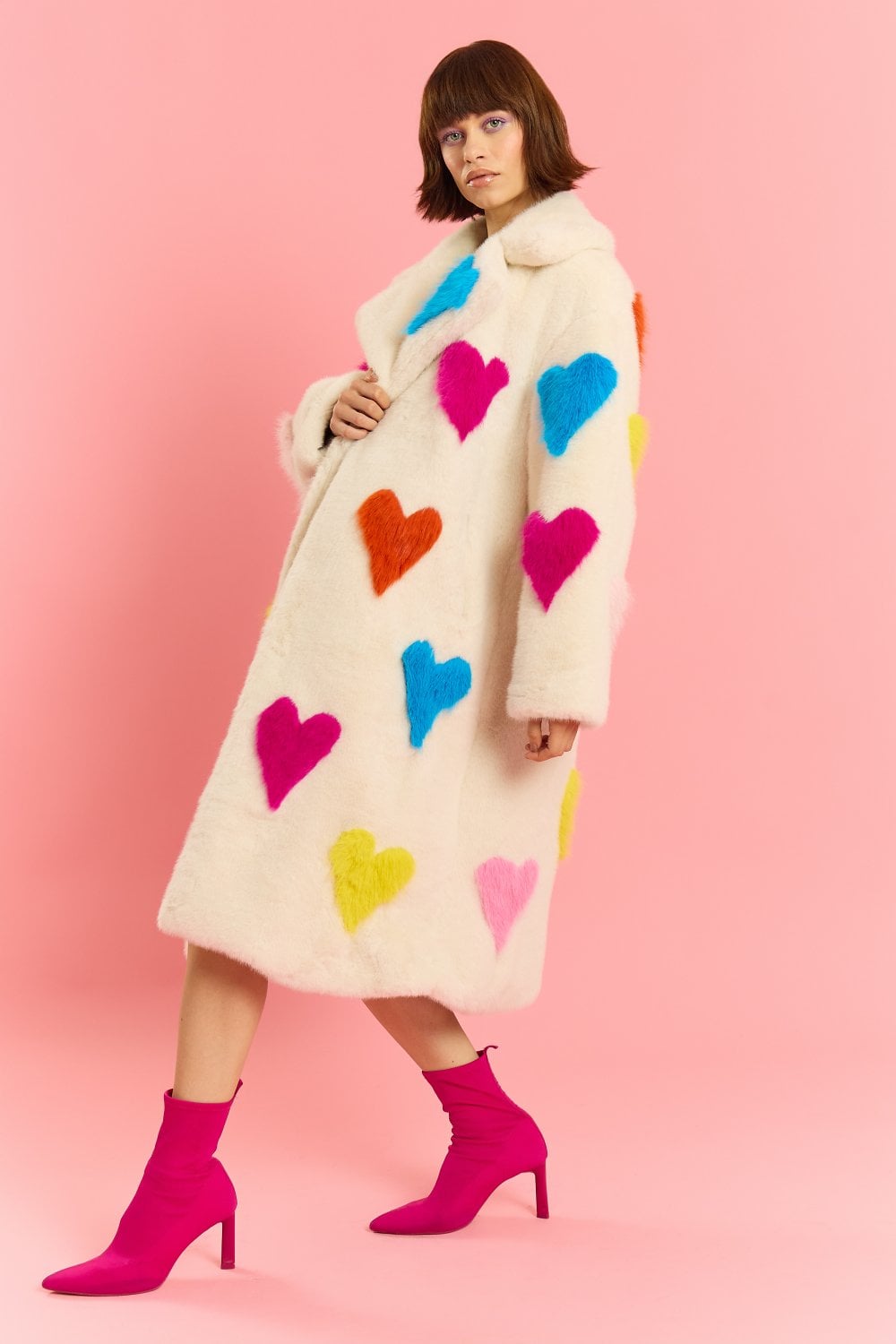 A person poses against a pink background wearing a long, sustainable faux fur coat with a colourful love heart pattern and bright pink heeled boots.