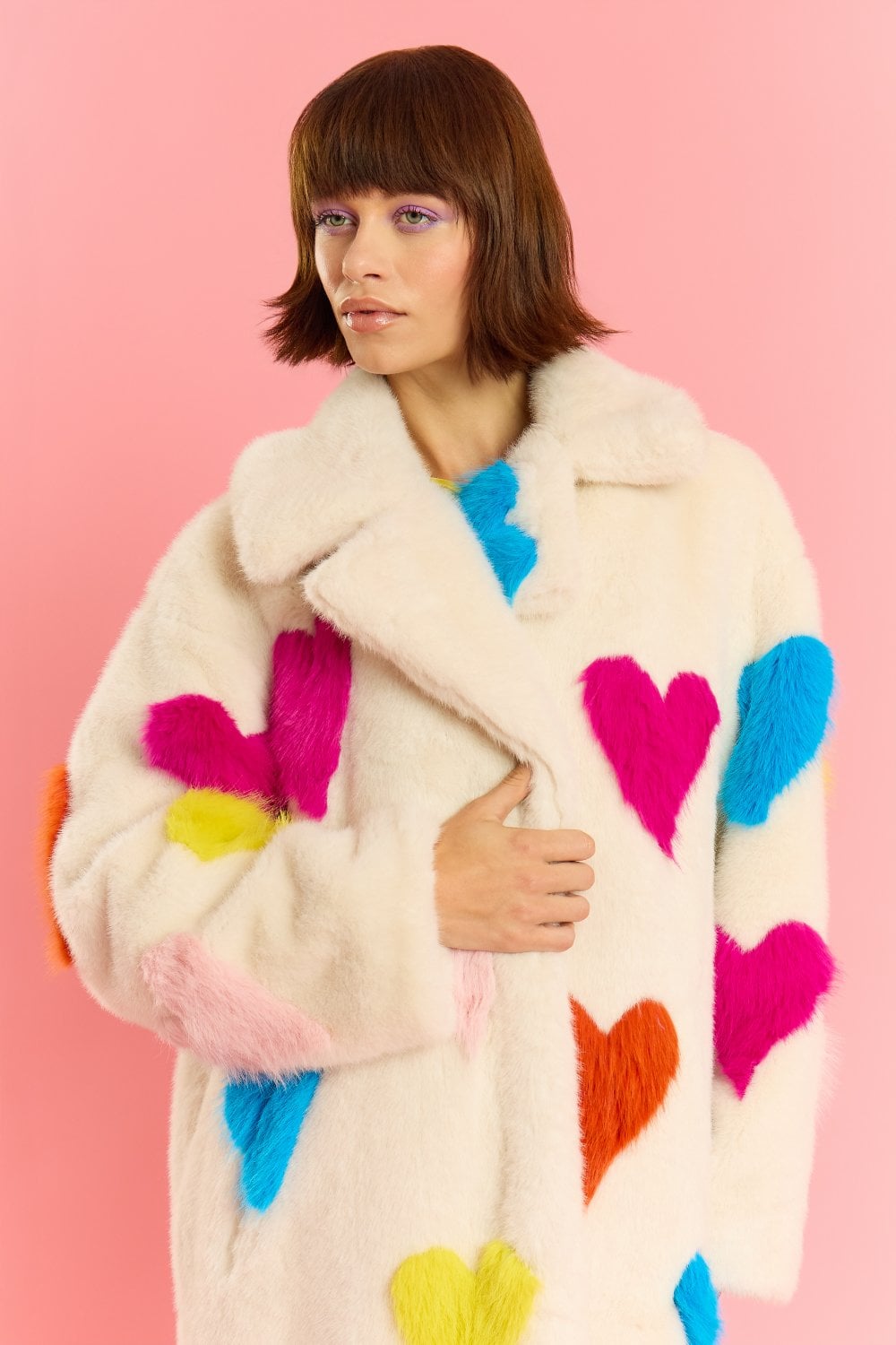 A person stands before a solid pink background, dressed in a cream-coloured love heart coat crafted from sustainable faux fur.