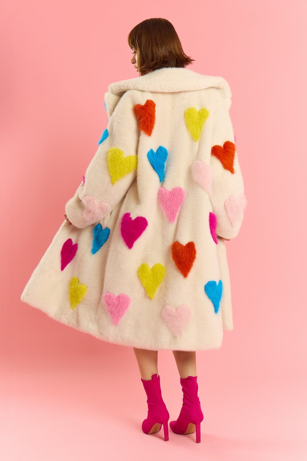 A person stands against a pink background wearing a long white sustainable faux fur coat with colourful love heart design and hot pink heeled boots. The person is facing away from the camera.