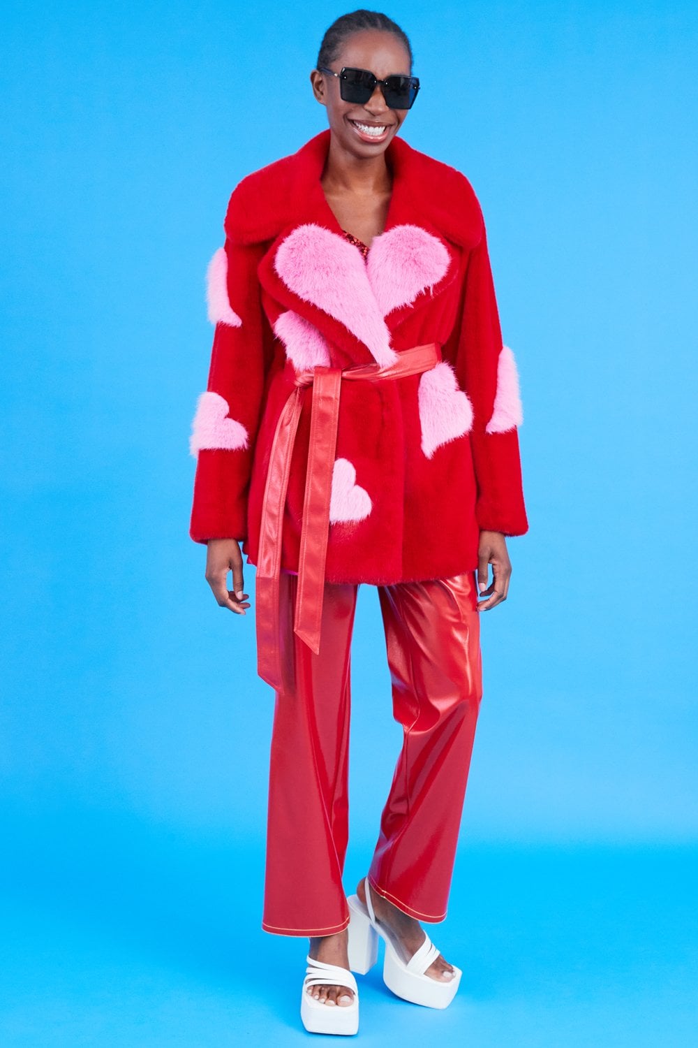 Person wearing an oversized belted Eco Bamboo Faux Fur Coat with a red and pink heart pattern, red vinyl trousers, white platform sandals, and sunglasses, standing against a blue background.
