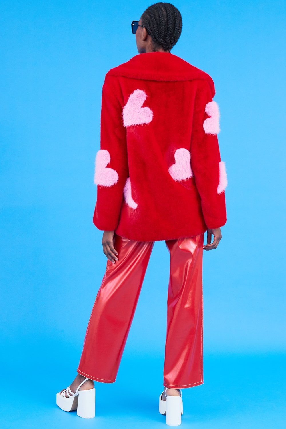 Person wearing a sustainable faux fur coat with pink heart patches, red shiny trousers, white platform shoes, and sunglasses stands confidently against a blue background.