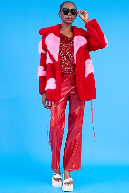 A person in an oversized belted sustainable faux fur coat, red sequinned top, red vinyl trousers, white platform sandals, and black sunglasses stands against a blue background.