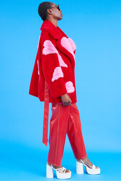 A person dressed in an oversized belted red coat adorned with pink heart patterns, red trousers, white platform sandals, and sunglasses, standing against a blue background.