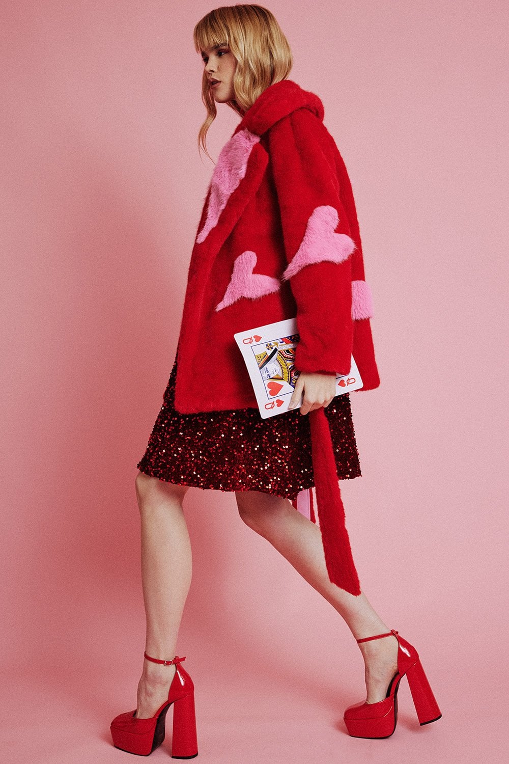 A woman wearing a sustainable faux fur coat with heart patterns, a red sequin skirt, red platform heels, and holding a Queen of Hearts clutch poses against a pink backdrop.