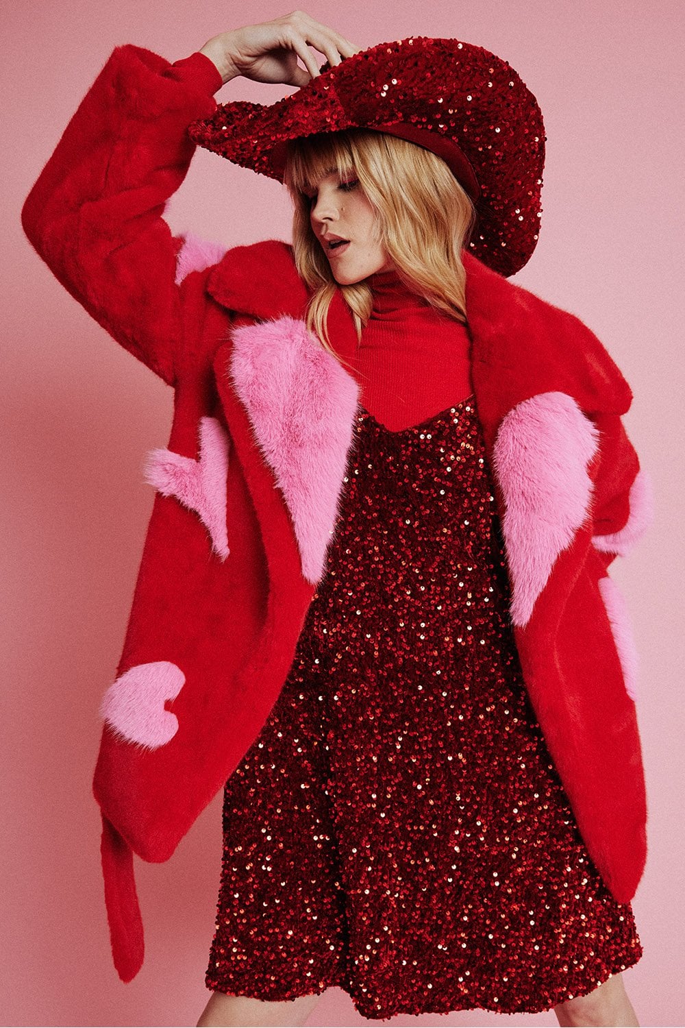 A person dressed in a red sequin dress, red polo neck, oversized belted Eco Bamboo Faux Fur Coat adorned with heart shapes, and a red sequin cowboy hat stands before a pink background.