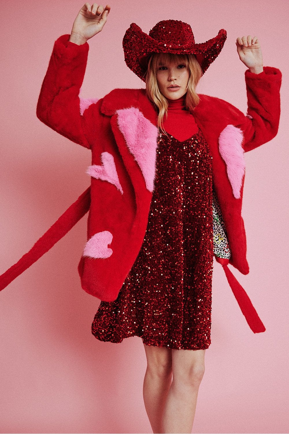 A person poses against a pink background wearing a red cowboy hat, a red sequin dress, and an Eco Bamboo faux fur coat adorned with pink heart patterns.
