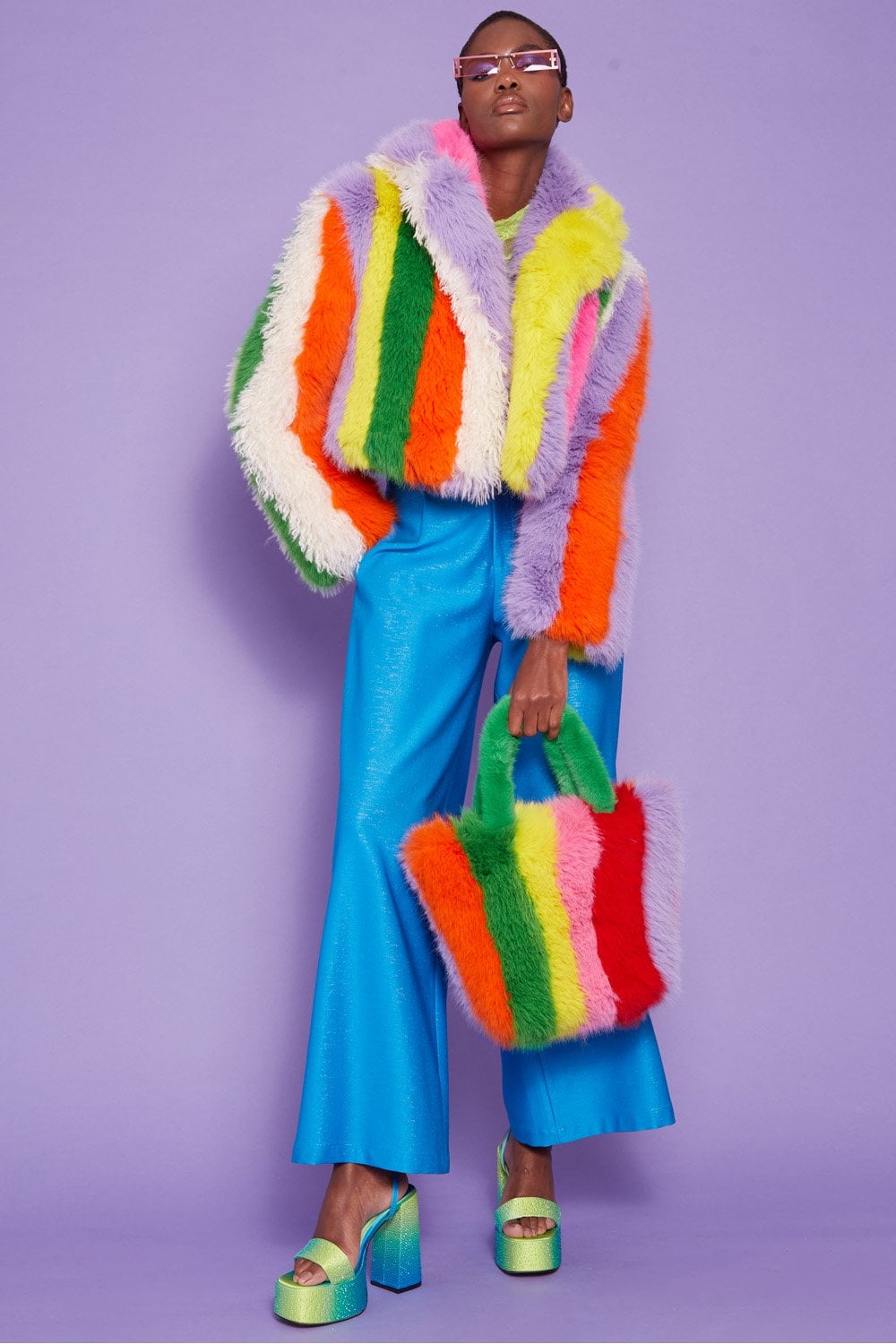 A person poses against a purple background wearing a rainbow-pattern faux fur coat, bright blue wide-leg trousers, green platform heels, and holding a matching striped faux fur bag.