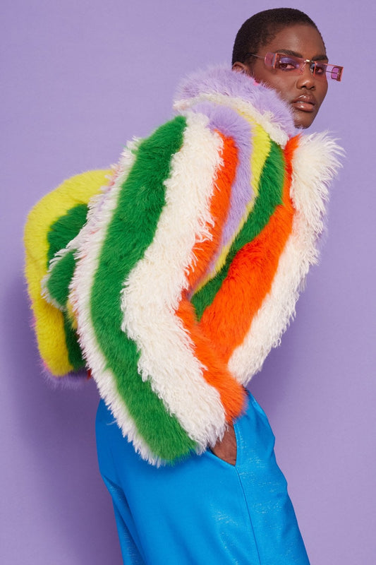Person wearing bright blue trousers and a rainbow-patterned coat—a multicoloured, striped faux fur jacket featuring green, orange, yellow, white, and purple—standing against a lavender background.