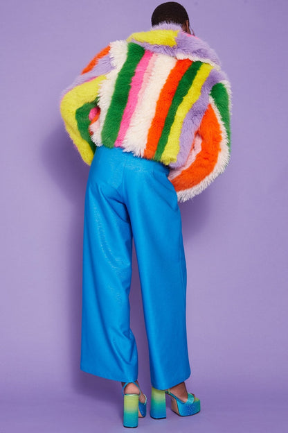 A person stands against a purple background wearing bright blue trousers, blue platform heels, and a faux fur coat with a colourful striped design, their back to the camera.