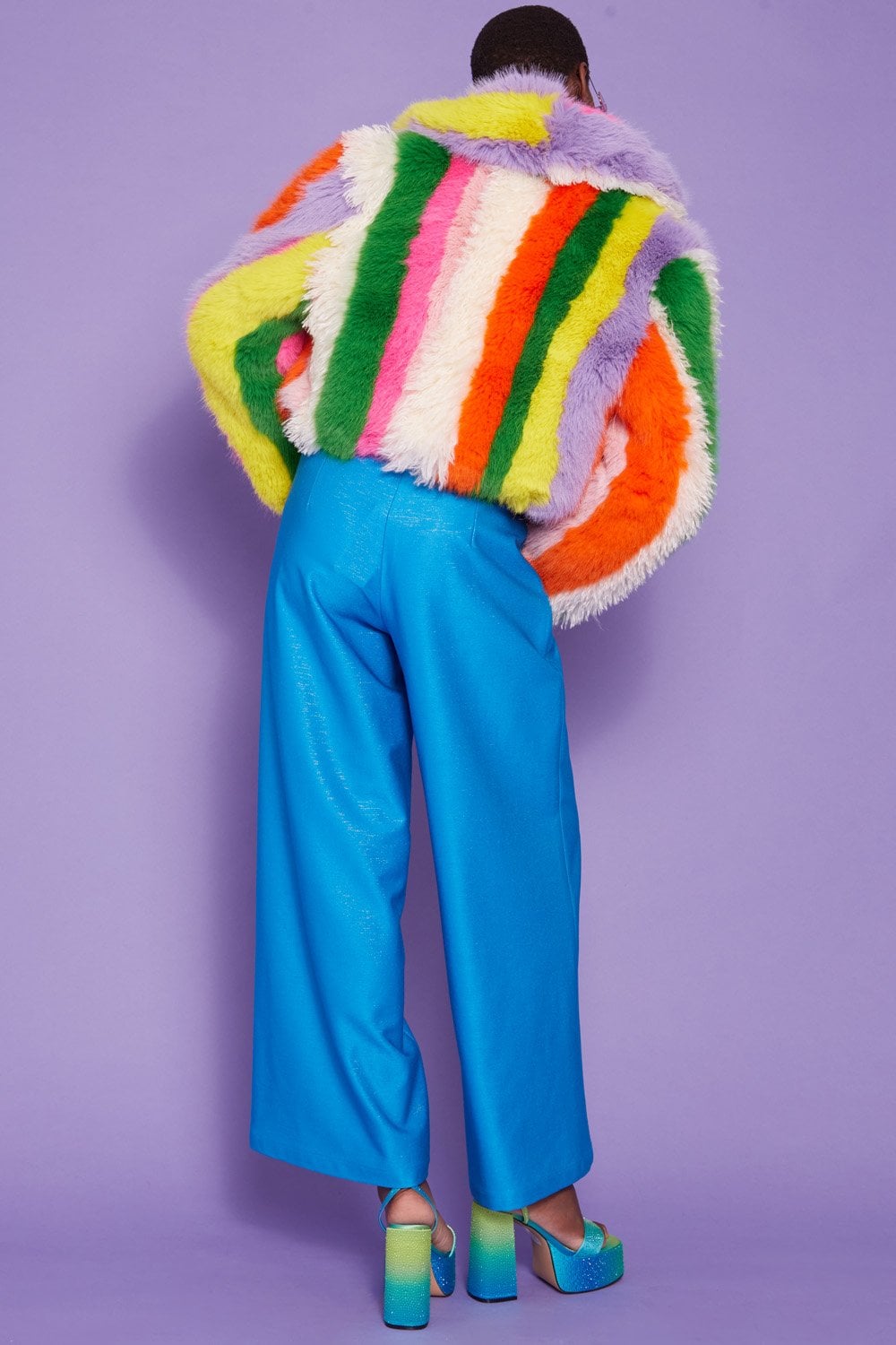 Person standing against a purple background, wearing bright blue wide-leg trousers, blue platform shoes, and a rainbow-pattern faux fur coat with colourful vertical stripes.