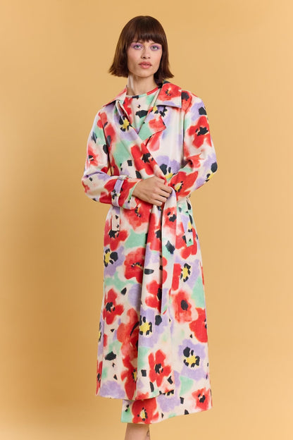 A woman stands against a beige background, wearing a long, colourful floral trench coat featuring red, purple, yellow and green patterns.