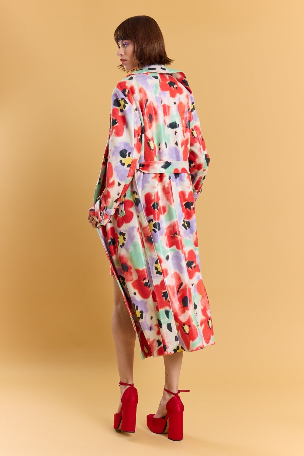 A person stands with their back to the viewer, dressed in a long, colourful floral trench coat with a belted waist and red high-heeled shoes, set against a plain beige background.