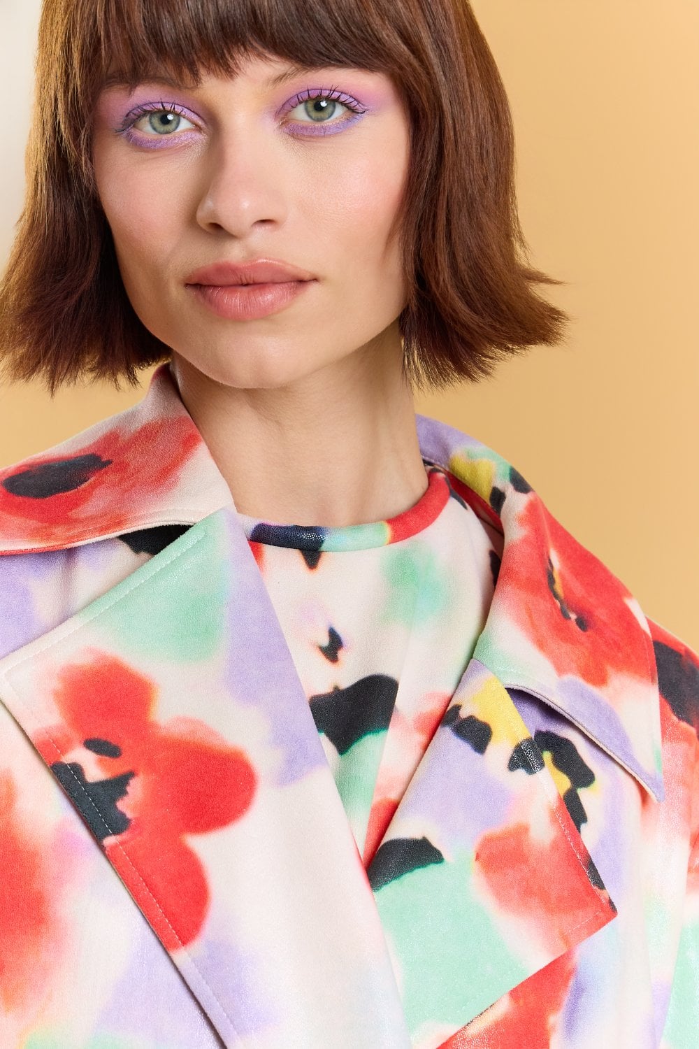 Woman with short brown hair and lilac eyeshadow wears a colourful floral trench coat and top. The background is a solid, warm beige colour.