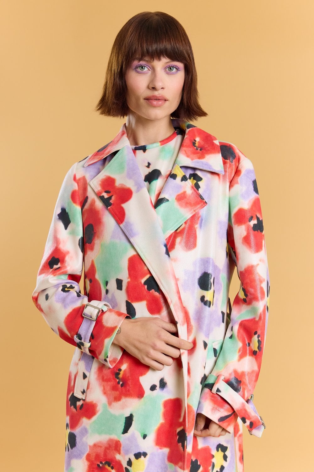 A woman with short brown hair wears a colourful, abstract faux suede floral trench coat and poses against a plain beige background.