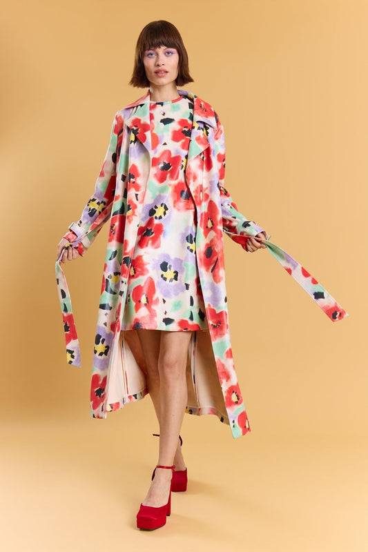 A woman stands against a tan background wearing a floral trench coat and matching dress with red, green, black, and yellow patterns. She finishes her look with red platform heels.
