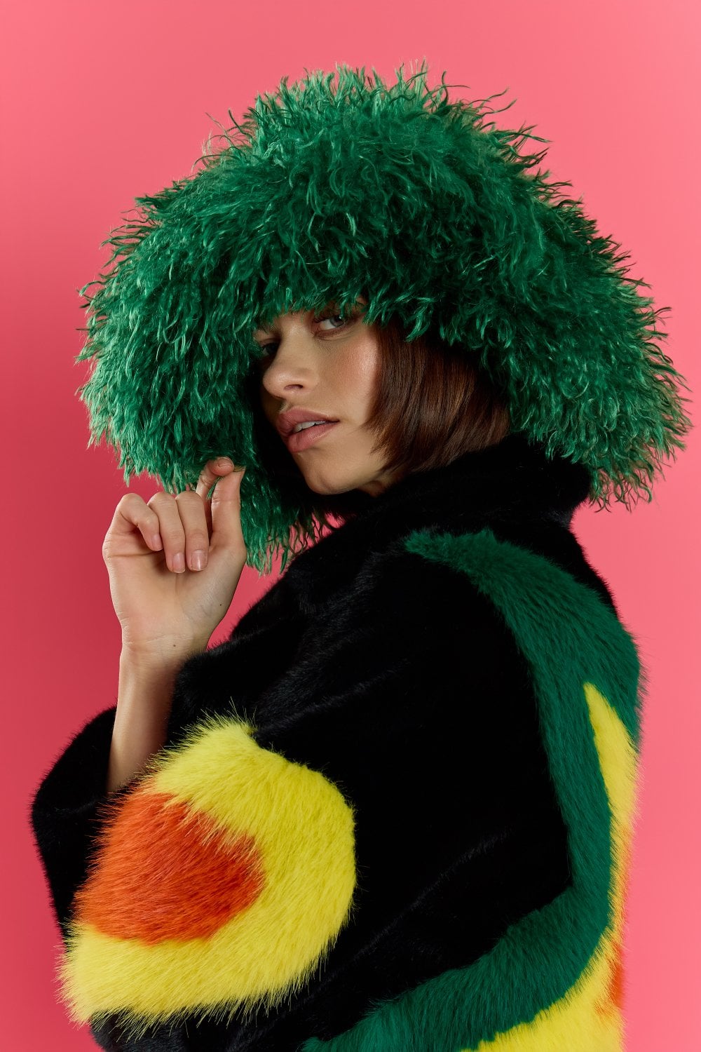 Woman wearing a large, fluffy green oversized bamboo fur hat and a colourful, furry coat with yellow and orange patches, posing against a pink background.