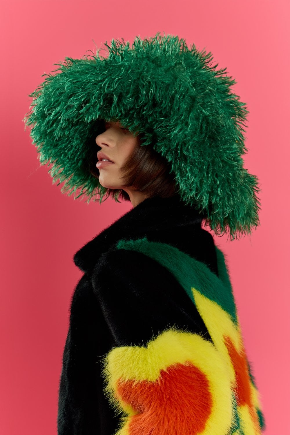 Person wearing an oversized green bamboo fur hat and a black coat featuring yellow and orange fur patterns, posed against a solid pink background.