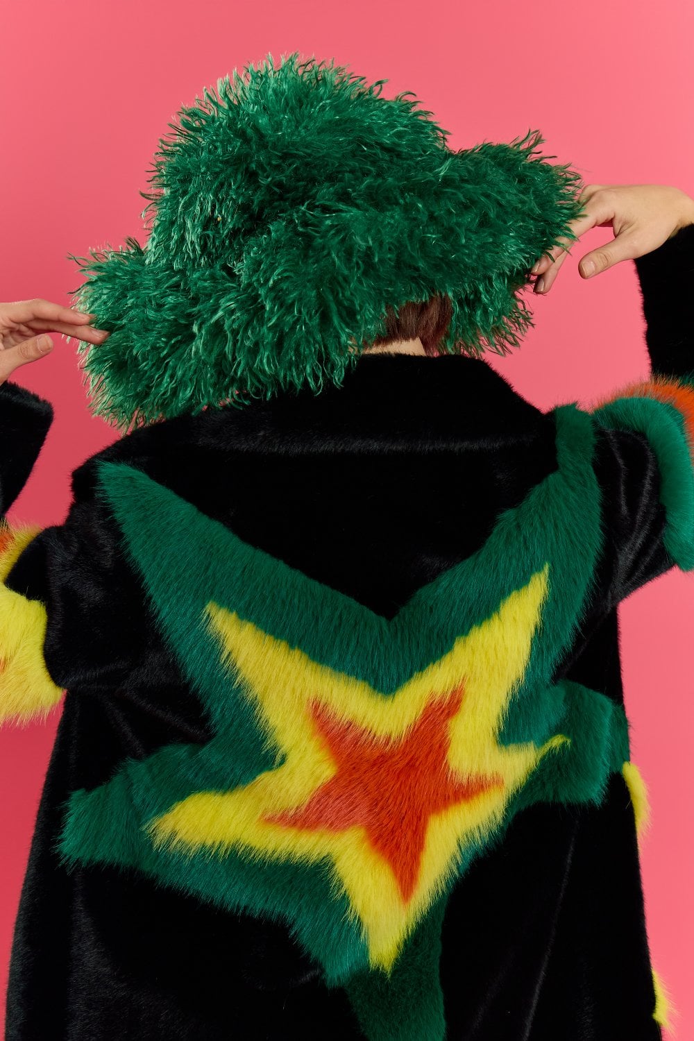 Person with their back to the viewer, wearing a large green bamboo fur hat and a black faux fur coat featuring bold yellow, green, and red star designs, standing before a pink backdrop.