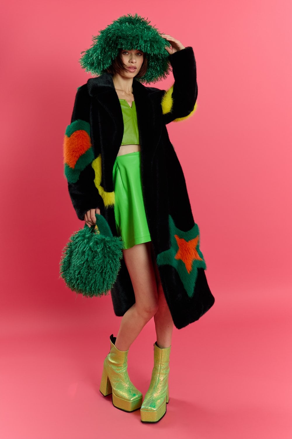A person dressed in a black coat with colourful star patterns, a green skirt and top, a luxury eco-friendly hat, furry green bag, and shiny green platform boots stands against a pink background.