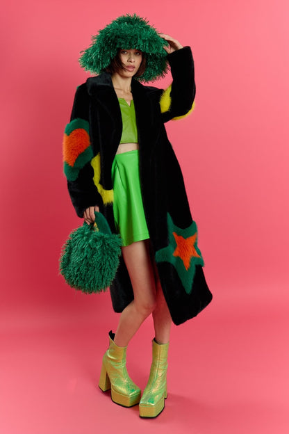 A person dressed in a black coat with colourful star patterns, a green skirt and top, a luxury eco-friendly hat, furry green bag, and shiny green platform boots stands against a pink background.
