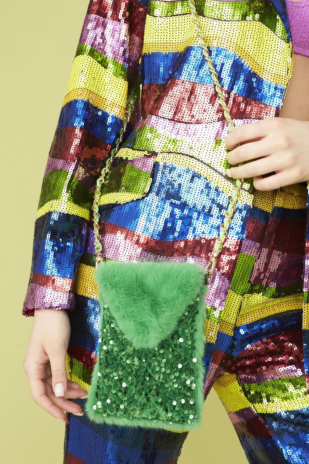 A person in a colourful, sequinned outfit holds a Stylish Phone Bag—a small green faux fur and sequin handbag with a gold chain strap—set against a vibrant yellow background.
