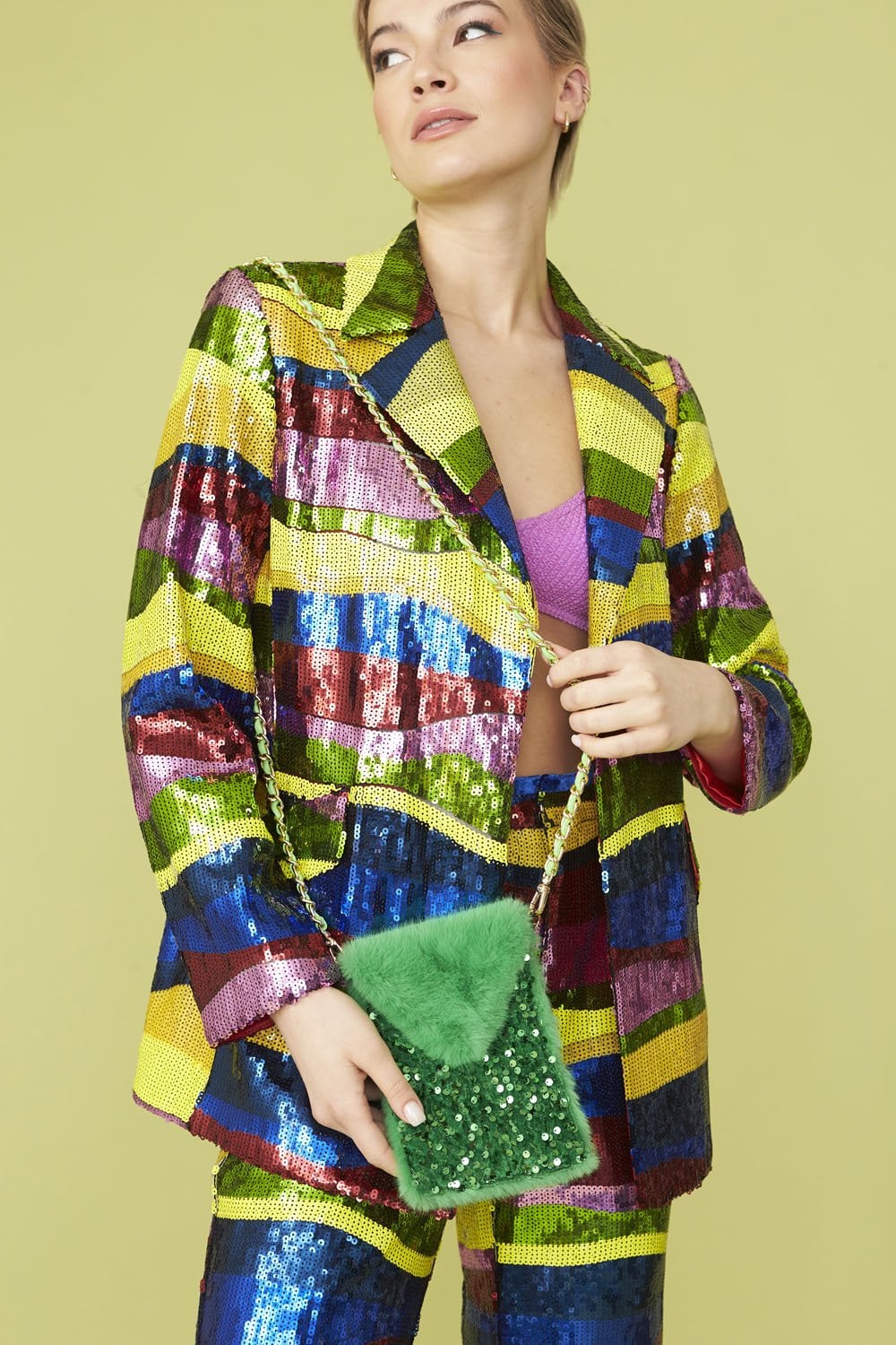 Person wearing a colourful sequin suit and purple bralette holds a green faux fur phone holder adorned with beads, posing against a light green background.
