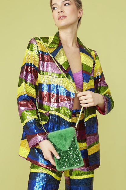 Person wearing a colourful sequin suit and purple bralette holds a green faux fur phone holder adorned with beads, posing against a light green background.
