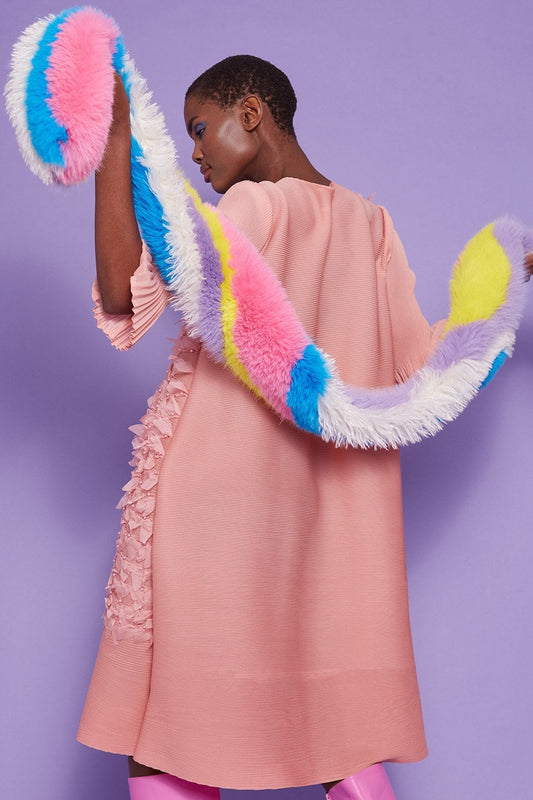 A person in a pale pink dress with frill details holds a long, sustainable faux fur scarf against a purple background.