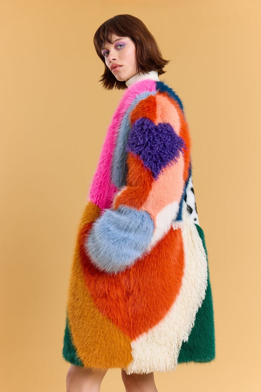 A person stands against a tan background, wearing a handmade faux fur coat featuring striking geometric patterns in pink, purple, blue, orange, green and white.