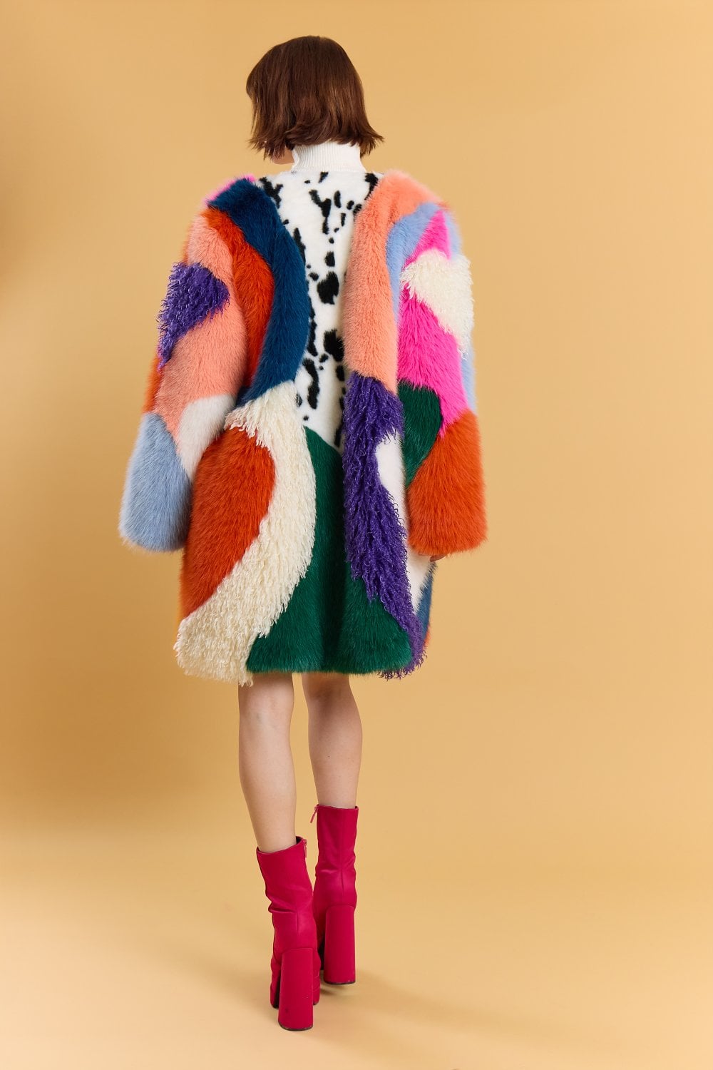 Person standing with their back to the camera, wearing a colourful, textured handmade faux fur coat featuring abstract patterns and hot pink platform boots, set against a plain beige background.