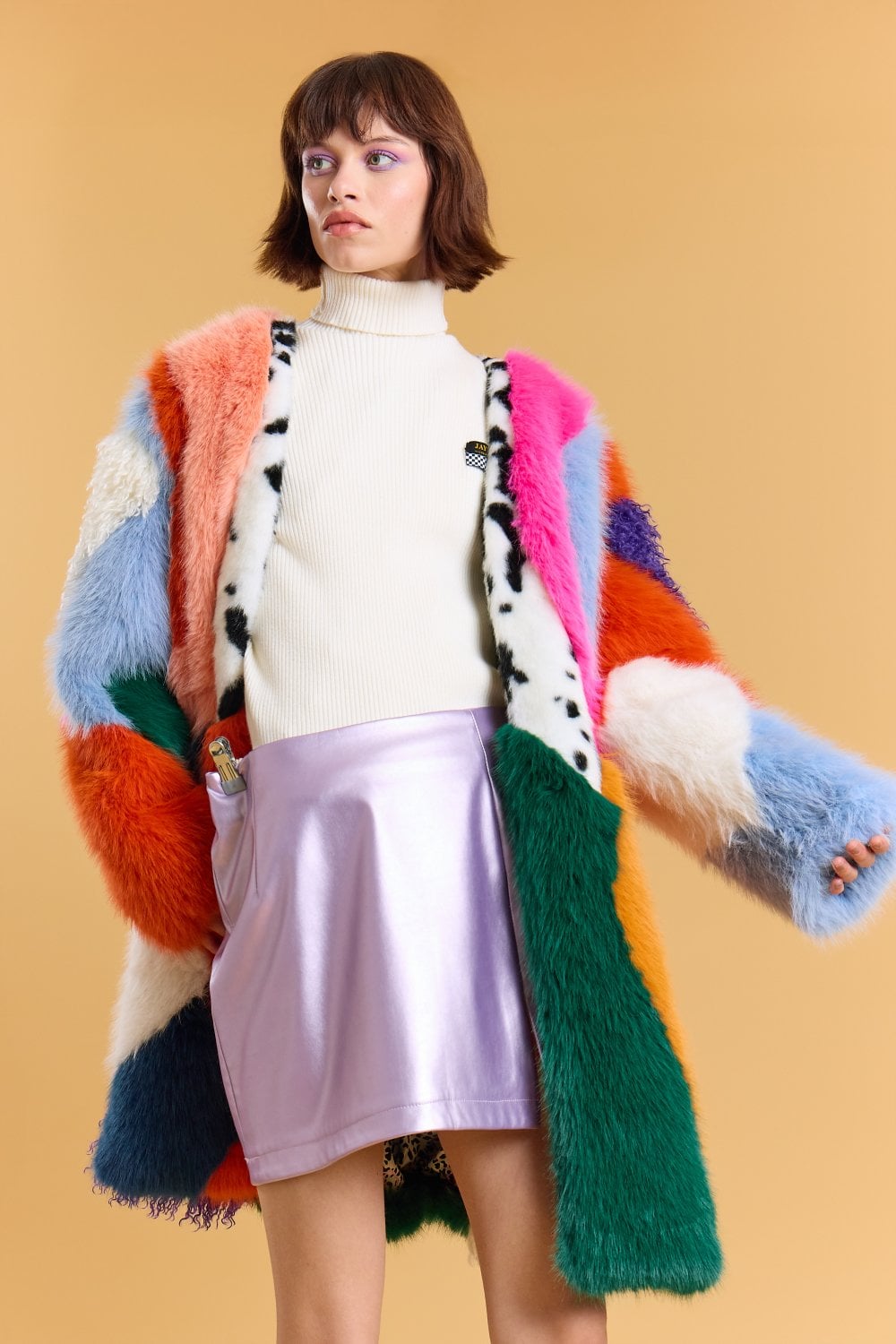 Woman in a colourful handmade faux fur coat, white polo neck, and shiny lilac mini skirt, standing against a plain tan backdrop.