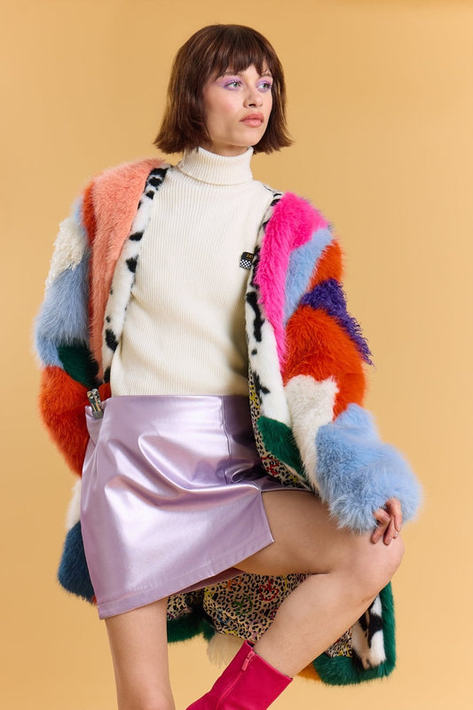 A woman poses in a white polo neck, metallic lavender skirt, and a colourful sustainable faux fur coat with patchwork patterns, standing against a beige background.