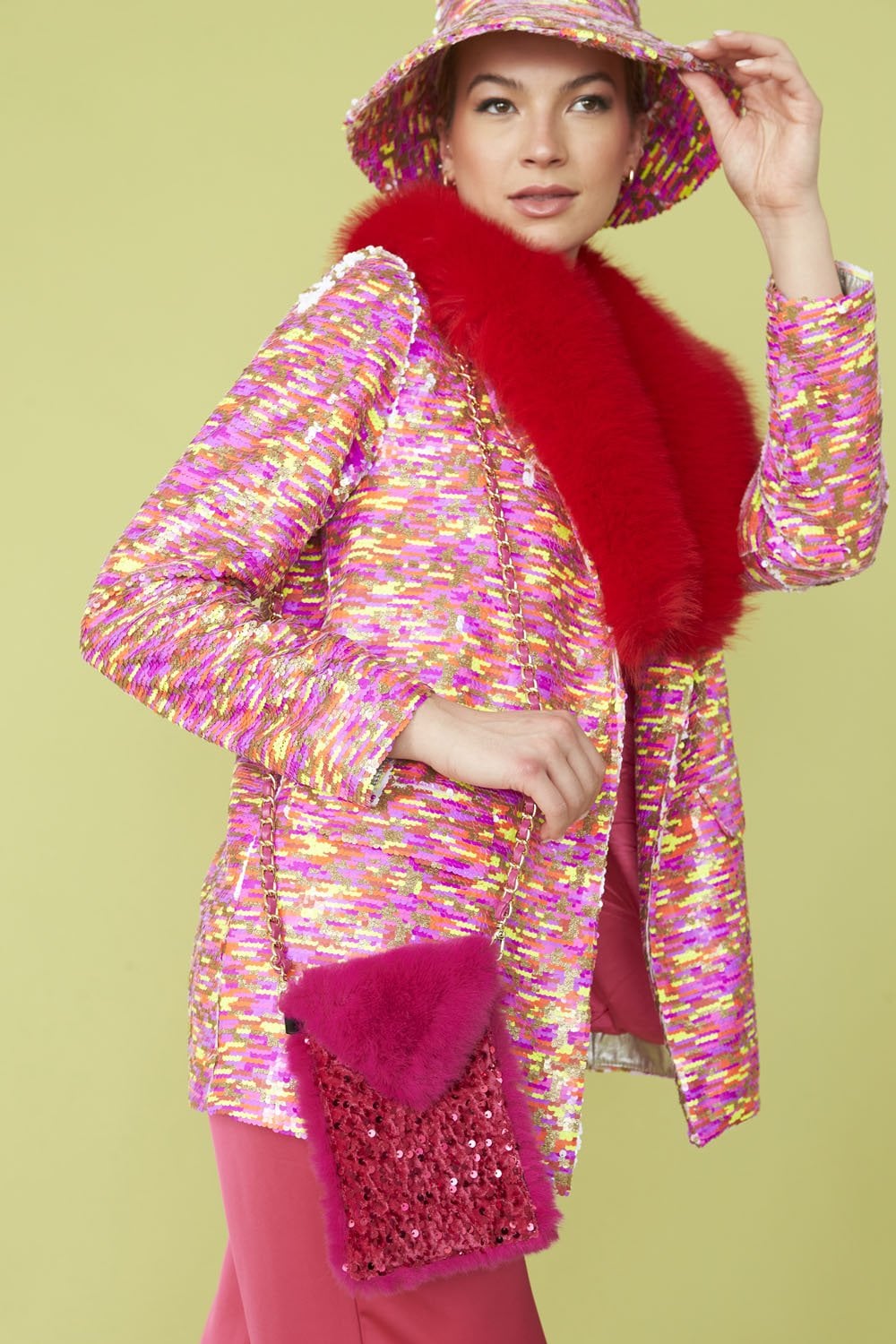 A woman wearing a colourful, patterned jacket, red fur stole, matching hat, and holding a sequinned phone holder bag against a pale yellow background.