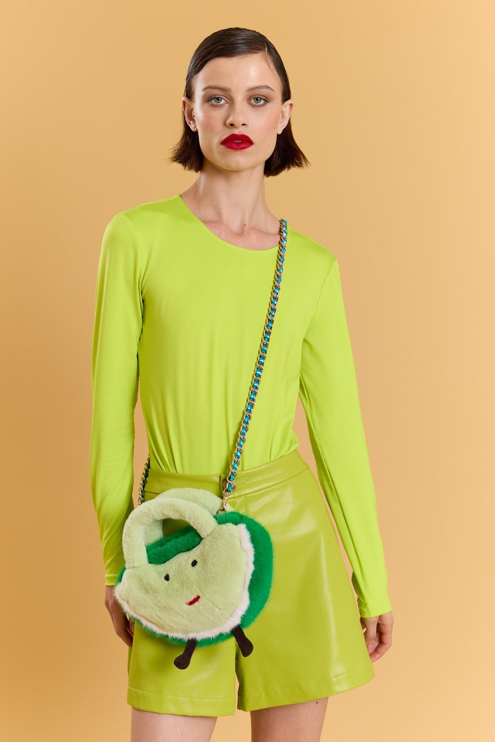 Woman in a neon green long-sleeved top and matching shorts, accessorised with a faux fur bag featuring a cartoon face, stands against a solid peach-coloured background.