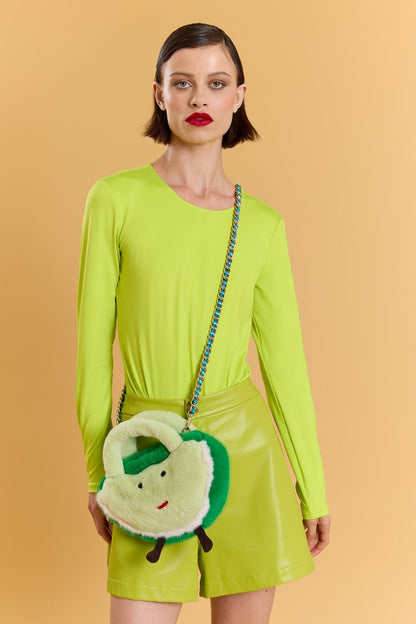 Woman in a neon green long-sleeved top and matching shorts, accessorised with a faux fur bag featuring a cartoon face, stands against a solid peach-coloured background.