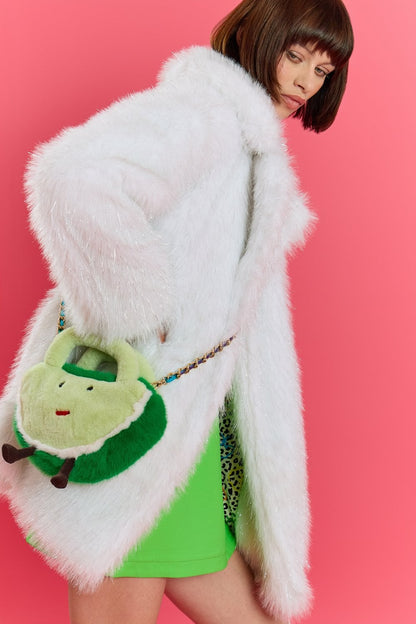 Woman in a white faux fur coat and green dress stands against a pink background, holding a plush green watermelon bamboo bag shaped like a smiling fruit—a playful nod to sustainable fashion.