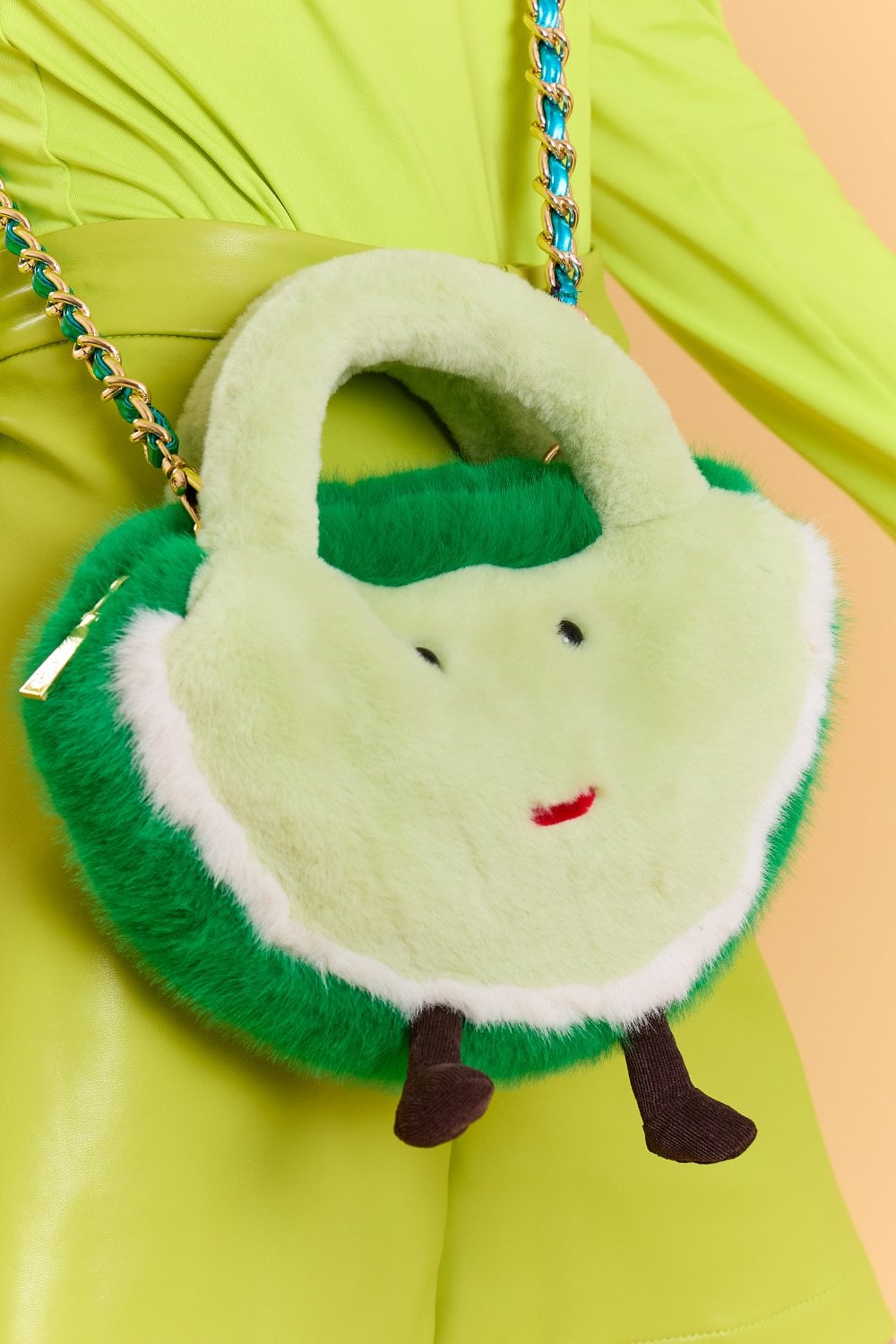 A person dressed in bright green clothing holds a plush, faux fur bag featuring a smiling face, short handles, chain strap, and small legs—playfully highlighting sustainable fashion.