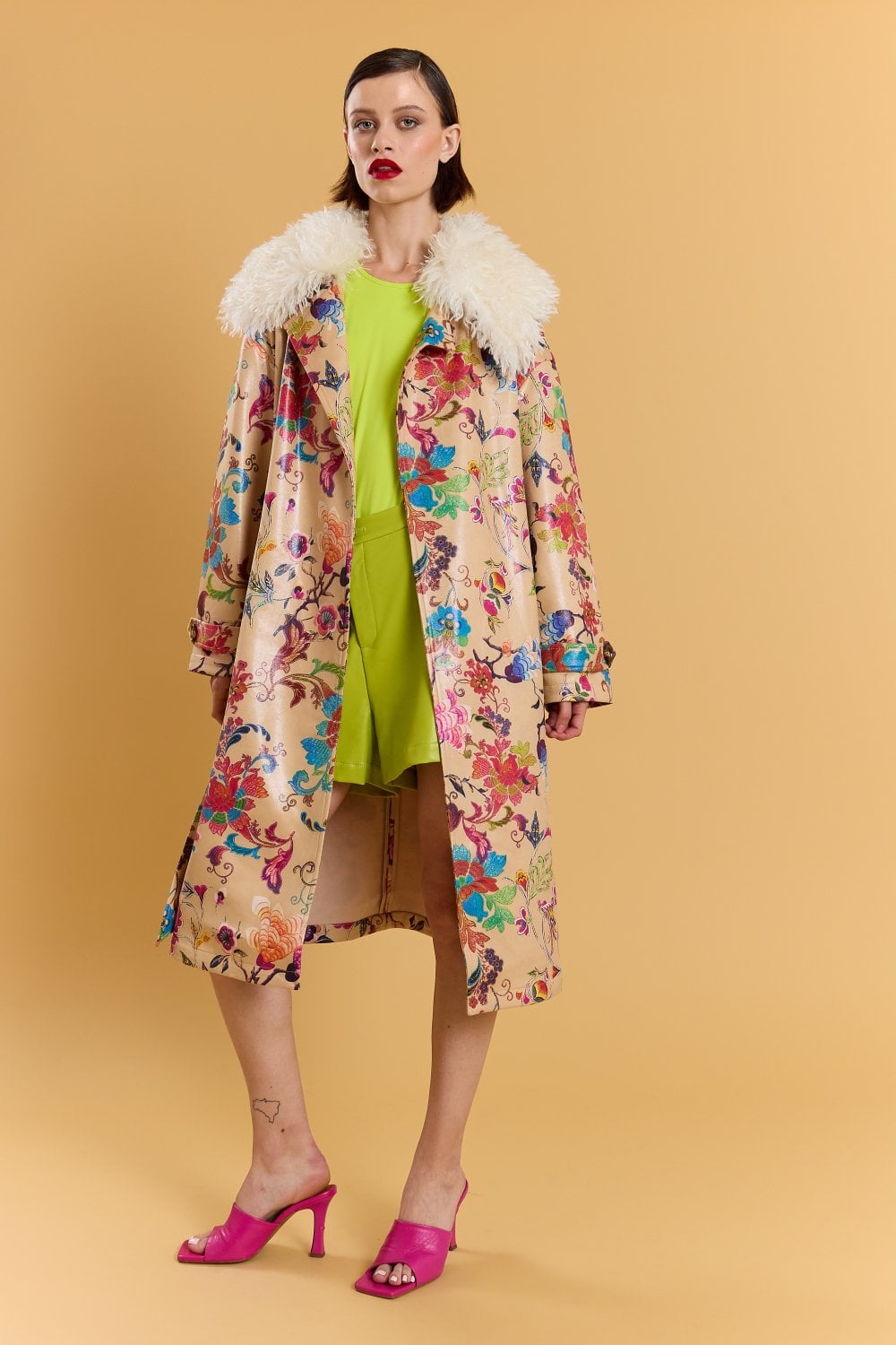 A woman stands against a beige background wearing an embroidered trench coat with a faux Mongolian fur collar, layered over a neon green dress and bright pink heeled sandals.