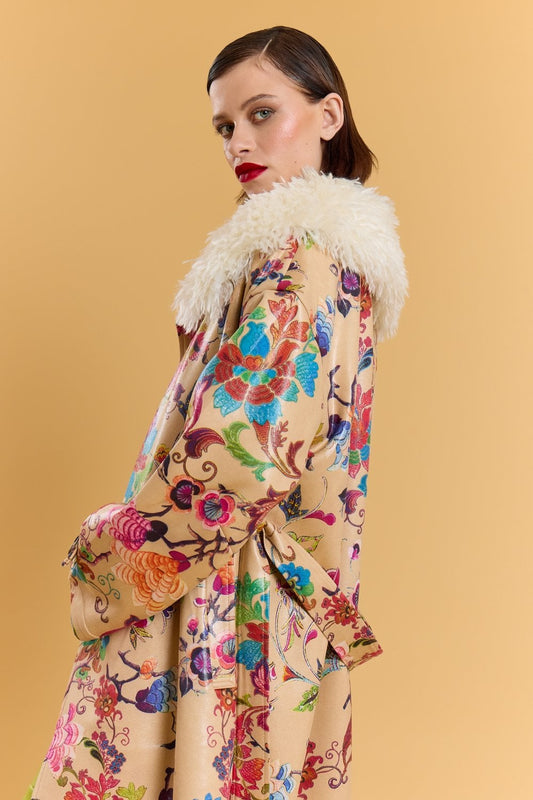 Person in an embroidered trench coat with a white faux Mongolian fur collar, standing before a plain beige background and glancing over their shoulder.