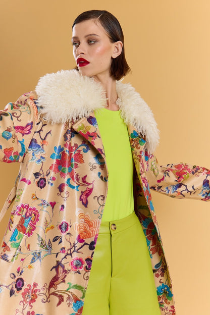 Person in an embroidered trench coat with a faux Mongolian fur collar over a vivid yellow-green outfit, standing before a plain beige background.