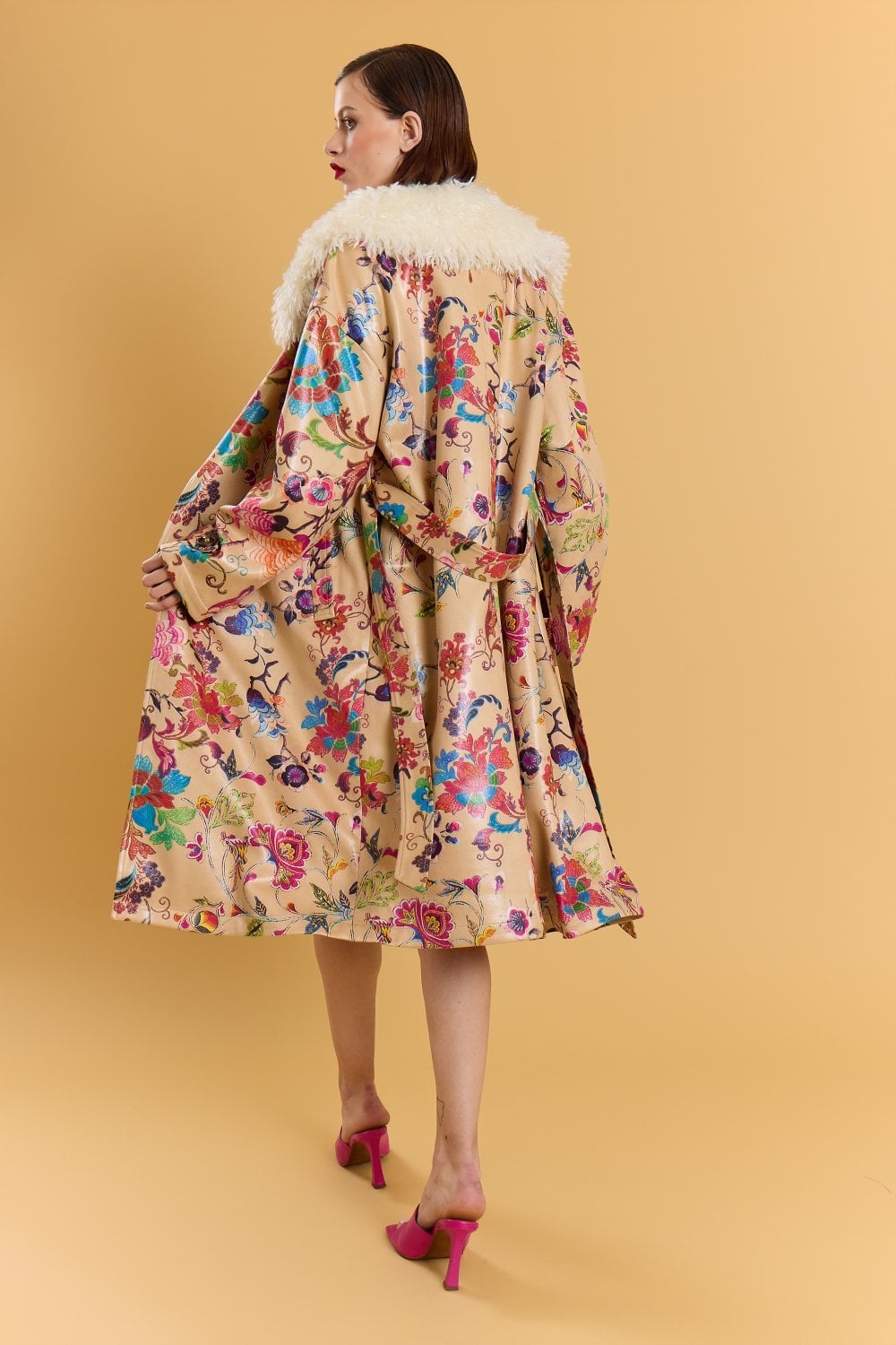 A woman stands against a beige background wearing a mocha faux suede trench coat with a floral pattern and faux Mongolian fur collar, paired with pink high heels, glancing over her shoulder.