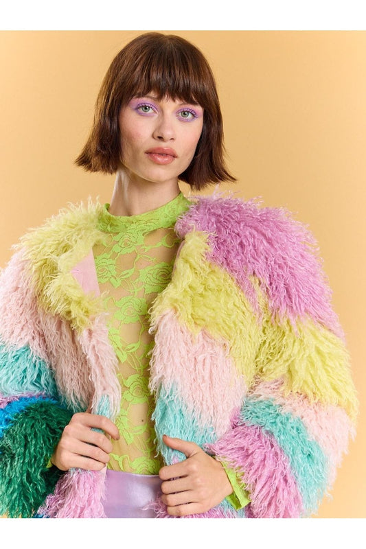 Woman with short brown hair wears a neon green lace top and a pastel rainbow faux fur coat in shades of pink, yellow, green, and blue, standing against a plain beige background.