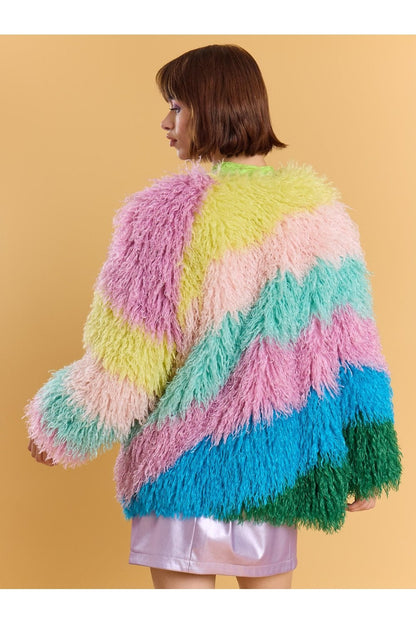 A person with short brown hair wears a pastel rainbow faux fur coat and a shiny light pink skirt, standing against a plain beige backdrop.